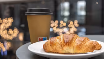 En croissant på en vit tallrik sitter bredvid en kaffekopp av brunt papper med svart lock. Bakgrunden har suddiga gyllene ljus, vilket skapar en mysig och inbjudande atmosfär.