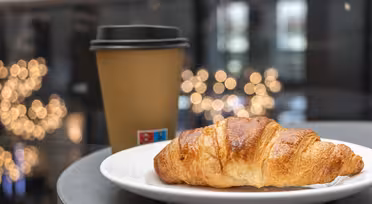 En croissant på en vit tallrik sitter bredvid en kaffekopp av brunt papper med svart lock. Bakgrunden har suddiga gyllene ljus, vilket skapar en mysig och inbjudande atmosfär.