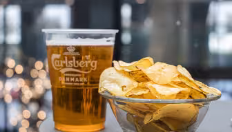En plastmugg med Carlsberg-öl står bredvid en glasskål med potatischips på ett bord. Bakgrunden är suddig med varm, mjuk belysning.
