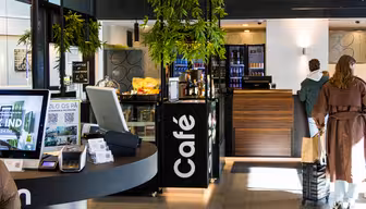Modern caféinteriör med digitala skärmar, snacks och en snygg disk. En person i brun rock och resväska står i kö. Vertikal "Café"-skylt och krukväxter förstärker den moderna designen. Ljus, välkomnande atmosfär.