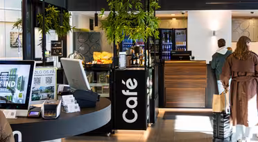 Modern caféinteriör med digitala skärmar, snacks och en snygg disk. En person i brun rock och resväska står i kö. Vertikal "Café"-skylt och krukväxter förstärker den moderna designen. Ljus, välkomnande atmosfär.