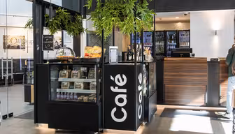 Modernt kafé med svartvitt färgschema, med en monter med bakverk, snacks och kaffepåsar. En menytavla listar kaffepriser. Det finns gröna växter ovanför, och en disk med kassaapparat finns i bakgrunden.