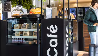 Ett mysigt café med utställning av bakverk, snacks och drycker. En person som bär en grön jacka drar en resväska. I caféets moderna interiör finns grönska och en svart skylt med "Café" skrivet på.