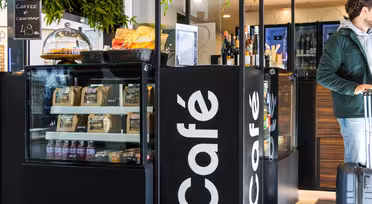 Ett mysigt café med utställning av bakverk, snacks och drycker. En person som bär en grön jacka drar en resväska. I caféets moderna interiör finns grönska och en svart skylt med "Café" skrivet på.