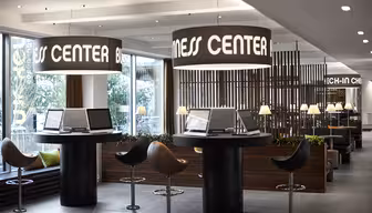 Modernt affärscenter med runda bord och datorer, upplyst av stora hängande lampor märkta "Business Center". I närheten förstärker eleganta snurrstolar och grönska den moderna inredningen. Naturligt ljus filtreras genom stora fönster.