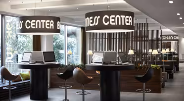 Modernt affärscenter med runda bord och datorer, upplyst av stora hängande lampor märkta "Business Center". I närheten förstärker eleganta snurrstolar och grönska den moderna inredningen. Naturligt ljus filtreras genom stora fönster.