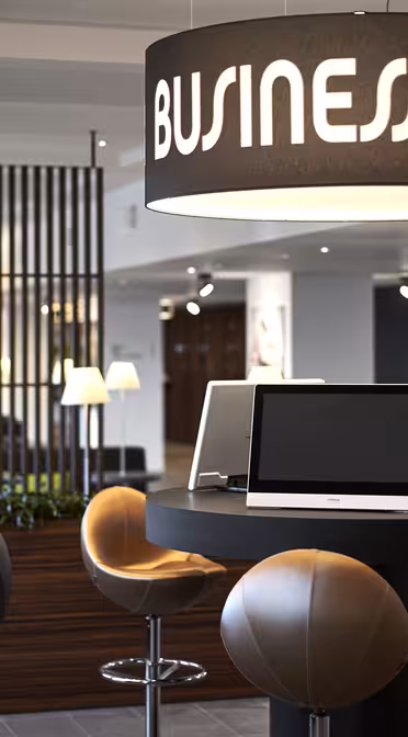 Modern arbetsyta med höga bord och stolar under stora "Business Center"-skyltar. Flera datorer finns tillgängliga på borden. Området har elegant belysning och inredning, med trädetaljer och växter i bakgrunden.