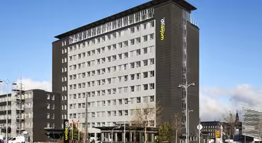Ett modernt hotell i flera våningar med mörkgrå exteriör och stora fönster, döpt till "Wakeup" med en gul logotyp. Byggnaden är omgiven av en parkeringsplats, gatlyktor och några parkerade bilar. Himlen är klar med några moln.