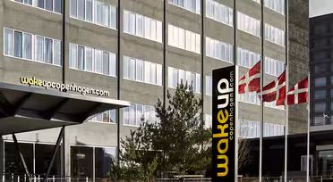 Bilden visar utsidan av Wakeup Copenhagen hotel med en stor skylt. Byggnaden är modern med många fönster. Tre danska flaggor vajar bredvid byggnaden. Träd och enkel landskapsarkitektur syns framför.