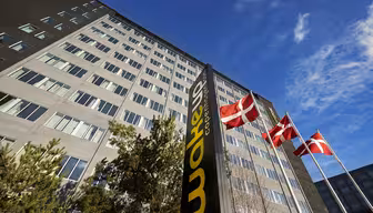 En hög byggnad med flera fönster ses från en låg vinkel. Tre danska flaggor vajar på stolpar framför den. En svart skylt med gula bokstäver lyder "Wakeup Copenhagen". Himlen är klar och blå.