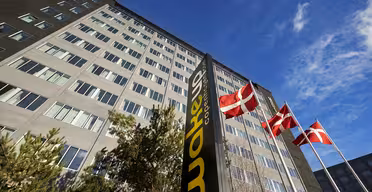 En hög byggnad med flera fönster ses från en låg vinkel. Tre danska flaggor vajar på stolpar framför den. En svart skylt med gula bokstäver lyder "Wakeup Copenhagen". Himlen är klar och blå.