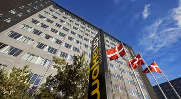 En hög byggnad med flera fönster ses från en låg vinkel. Tre danska flaggor vajar på stolpar framför den. En svart skylt med gula bokstäver lyder "Wakeup Copenhagen". Himlen är klar och blå.