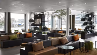 Modern, rymlig lobby med stora fönster. Har mörkgrå soffor med orange och beige kuddar, runda bord och grönska på hyllor. Rikligt naturligt ljus skapar en inbjudande atmosfär.