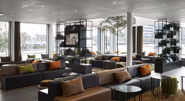 Modern, rymlig lobby med stora fönster. Har mörkgrå soffor med orange och beige kuddar, runda bord och grönska på hyllor. Rikligt naturligt ljus skapar en inbjudande atmosfär.