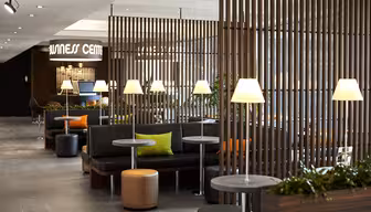 Modern lounge med svarta sittplatser, trädetaljer och mjuk belysning. Flera små bord med lampor och gröna kuddar ger en touch av färg. En skylt "Hälsocenter" syns i bakgrunden.