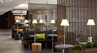 Modern lounge med svarta sittplatser, trädetaljer och mjuk belysning. Flera små bord med lampor och gröna kuddar ger en touch av färg. En skylt "Hälsocenter" syns i bakgrunden.
