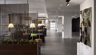 Modern hotellobby med eleganta träavdelare, krukväxter och bekväma sittplatser. En väggdetalj med spegelpaneler pryder sidan. Mjuk belysning skapar en inbjudande atmosfär. Ett incheckningsområde syns i bakgrunden.