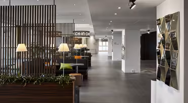 Modern hotellobby med eleganta träavdelare, krukväxter och bekväma sittplatser. En väggdetalj med spegelpaneler pryder sidan. Mjuk belysning skapar en inbjudande atmosfär. Ett incheckningsområde syns i bakgrunden.