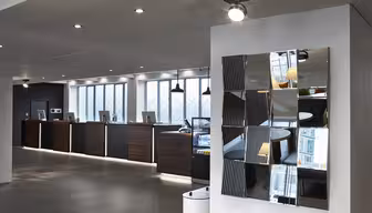 Bilden visar en modern hotellreception med en rad träreceptioner utrustade med datorer. Ovanför varje skrivbord hänger en taklampa. På den vita väggen till höger finns en dekorativ spegel sammansatt av fragmenterade paneler.