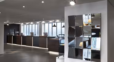 Bilden visar en modern hotellreception med en rad träreceptioner utrustade med datorer. Ovanför varje skrivbord hänger en taklampa. På den vita väggen till höger finns en dekorativ spegel sammansatt av fragmenterade paneler.