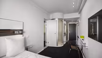 Modernt hotellrum med minimalistisk design, med en vit säng, glasbord och en öppen cylindrisk dusch med en genomskinlig dörr. Rummet är ljust, med rena linjer och en elegant, modern estetik.