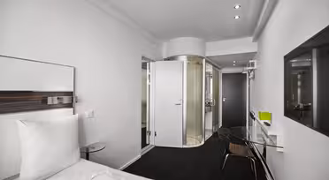 Modernt hotellrum med minimalistisk design, med en vit säng, glasbord och en öppen cylindrisk dusch med en genomskinlig dörr. Rummet är ljust, med rena linjer och en elegant, modern estetik.