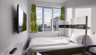 Ett litet, modernt hotellrum har en snyggt bäddad säng med vita sängkläder och gröna, mönstrade gardiner. En väggmonterad TV vetter mot sängen. Fönstret erbjuder utsikt över stadsbilden och släpper in naturligt ljus. En fjärrkontroll och skrivbordsstol är synliga.