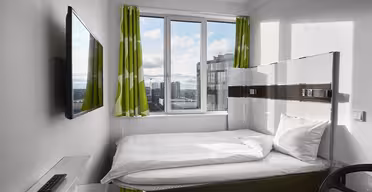 Ett litet, modernt hotellrum har en snyggt bäddad säng med vita sängkläder och gröna, mönstrade gardiner. En väggmonterad TV vetter mot sängen. Fönstret erbjuder utsikt över stadsbilden och släpper in naturligt ljus. En fjärrkontroll och skrivbordsstol är synliga.