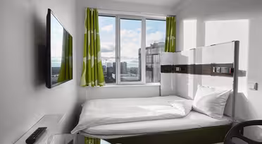 Ett litet, modernt hotellrum har en snyggt bäddad säng med vita sängkläder och gröna, mönstrade gardiner. En väggmonterad TV vetter mot sängen. Fönstret erbjuder utsikt över stadsbilden och släpper in naturligt ljus. En fjärrkontroll och skrivbordsstol är synliga.