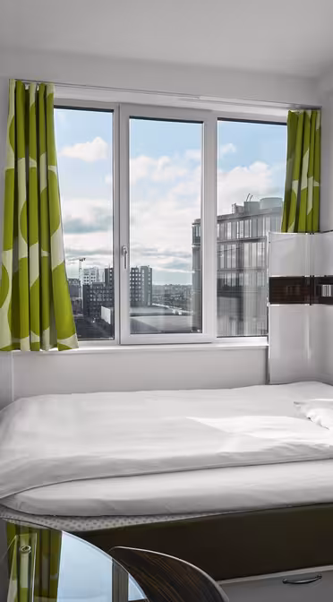 Ett litet, modernt hotellrum har en snyggt bäddad säng med vita sängkläder och gröna, mönstrade gardiner. En väggmonterad TV vetter mot sängen. Fönstret erbjuder utsikt över stadsbilden och släpper in naturligt ljus. En fjärrkontroll och skrivbordsstol är synliga.