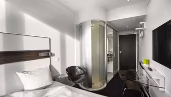 Modernt hotellrum med säng, skrivbord i glas, svart stol, väggmonterad TV och en cylindrisk duschvägg i glas. Rummet har minimalistisk inredning med vita väggar och eleganta möbler.