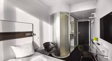 Modernt hotellrum med säng, skrivbord i glas, svart stol, väggmonterad TV och en cylindrisk duschvägg i glas. Rummet har minimalistisk inredning med vita väggar och eleganta möbler.