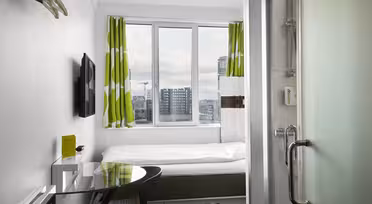Kompakt hotellrum med en enkelsäng bredvid ett fönster med gröna och vita gardiner. Rummet har en väggmonterad TV, ett litet skrivbord med stol och en glasdörr som leder till ett badrum. Stadsbilden syns genom fönstret.