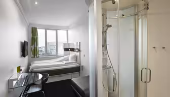Ett kompakt, modernt hotellrum med en dubbelsäng under ett fönster med gröna och vita gardiner. Det finns en väggmonterad TV, ett skrivbord i klarglas med en stol, en liten ottoman och en sluten dusch till höger. Inredningen är minimalistisk och elegant.