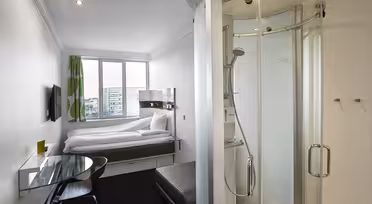 Ett kompakt, modernt hotellrum med en dubbelsäng under ett fönster med gröna och vita gardiner. Det finns en väggmonterad TV, ett skrivbord i klarglas med en stol, en liten ottoman och en sluten dusch till höger. Inredningen är minimalistisk och elegant.