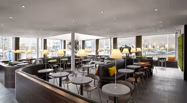Ett modernt, rymligt café med träbord och stolar och mysiga bås. Varm belysning från bordslampor skapar en välkomnande atmosfär. Stora fönster ger naturligt ljus och utsikt över staden utanför. Vertikal grönska pryder ena väggen.