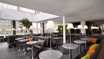 En modern caféinteriör med runda bord och vadderade sittplatser. Varje bord har en lampa, och gröna och orangea kuddar ger en färgklick. Stora fönster släpper in naturligt ljus, medan växter förstärker inredningen. Vita tak och neutrala toner skapar en lugn atmosfär.