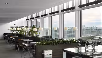 Modern matplats med stora fönster med utsikt över staden. Rader av bord och stolar kantar rummet, med växter och varm belysning skapar en välkomnande atmosfär. En bufféuppsättning med täckta rätter står i förgrunden.