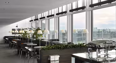 Modern matplats med stora fönster med utsikt över staden. Rader av bord och stolar kantar rummet, med växter och varm belysning skapar en välkomnande atmosfär. En bufféuppsättning med täckta rätter står i förgrunden.