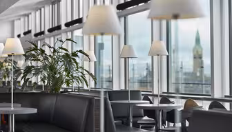 En modern lounge med stora fönster och naturligt ljus. Svarta vadderade sittplatser och runda bord med vita lampor är snyggt arrangerade. Gröna växter ger en touch av naturen. Ett torn syns genom fönstren i bakgrunden.