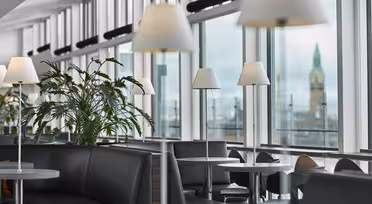 En modern lounge med stora fönster och naturligt ljus. Svarta vadderade sittplatser och runda bord med vita lampor är snyggt arrangerade. Gröna växter ger en touch av naturen. Ett torn syns genom fönstren i bakgrunden.