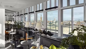En modern lounge med stora fönster med utsikt över staden. Utrymmet har bord med gröna stolar, svarta soffor och vita lampor. Krukväxter ger grönska, och en frukostmiljö med juice och ett bakverk står på ett bord.