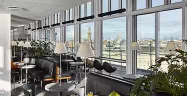 En modern lounge med stora fönster med utsikt över staden. Utrymmet har bord med gröna stolar, svarta soffor och vita lampor. Krukväxter ger grönska, och en frukostmiljö med juice och ett bakverk står på ett bord.