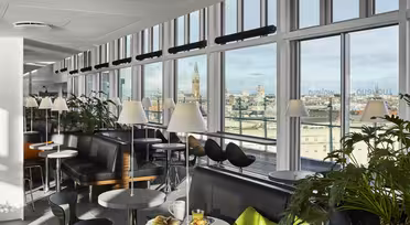 En modern lounge med stora fönster med utsikt över staden. Utrymmet har bord med gröna stolar, svarta soffor och vita lampor. Krukväxter ger grönska, och en frukostmiljö med juice och ett bakverk står på ett bord.