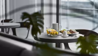 Ett modernt matbord med en skål med frukt, en kopp och en tallrik med ett bakverk och ett litet fat, delvis skymd av gröna växtblad. Bakgrunden har stora fönster med suddig utsikt utanför.