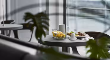 Ett modernt matbord med en skål med frukt, en kopp och en tallrik med ett bakverk och ett litet fat, delvis skymd av gröna växtblad. Bakgrunden har stora fönster med suddig utsikt utanför.