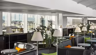 En modern lounge med stora fönster med utsikt över staden. Utrymmet har runda bord, vadderade sittplatser med gröna och orange kuddar och krukväxter. Vita lampor placeras på borden, vilket förstärker den rena, moderna designen.