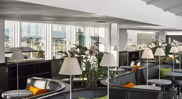 En modern lounge med stora fönster med utsikt över staden. Utrymmet har runda bord, vadderade sittplatser med gröna och orange kuddar och krukväxter. Vita lampor placeras på borden, vilket förstärker den rena, moderna designen.
