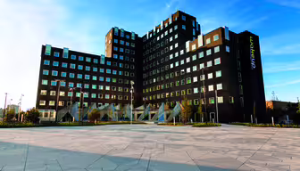 En modern hotellbyggnad i flera våningar med geometrisk design, med många fyrkantiga fönster och mörka ytterväggar. Det ligger mot en blå himmel, med en rymlig kaklad innergård i förgrunden.
