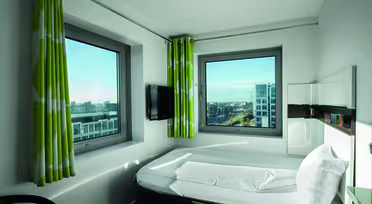 Ett hotellrum med en snyggt bäddad säng och två stora fönster med utsikt över stadsbilden. Väggarna är vita och det finns gröna gardiner med vita mönster. En väggmonterad TV finns bredvid sängen och solljus lyser upp rummet.